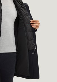 Cappotto nero isolato con interno trapuntato, tasche con zip e tessuto esterno testurizzato. Indumento interno mostrato parzialmente aperto.