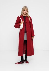 Long red coat with a notched collar and button closure, paired with a dark sweater and gray skirt. White handbag and red flats complete the outfit.