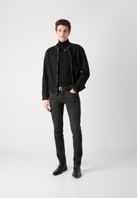 Black denim jacket with zip and snap closure, worn over a black turtleneck. Fitted black jeans and black leather shoes complete the outfit.