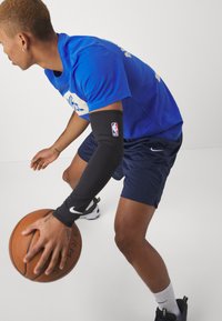 Nike športový outfit, ktorý obsahuje modré tričko, čiernu rukávovú vložku s logom a tmavomodré šortky. Subjekt driblí basketbal.