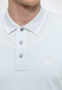 Polo bleu clair avec trois boutons et logo blanc "BOSS" sur la poitrine gauche, porté par une personne montrée du cou vers le bas.