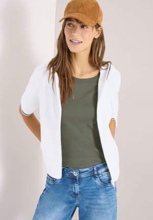 Junge Frau mit brainer Mütze, weißem kurzärmeligem Reißverschluss-Hoodie, grünem geripptem Tanktop und blauen Jeans, steht mit den Händen hinter dem Rücken.