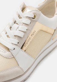 Gros plan d'une basket Michael Kors beige et dorée avec des lacets blancs et des détails en mesh sur un fond blanc.
