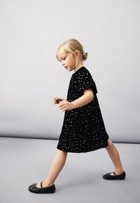 Mladá dívka s blond vlasy svázanými do drdolu, kráčející, oblečená v černých šatech s bílými puntíky a v černých balerínkách, na neutrálním pozadí.