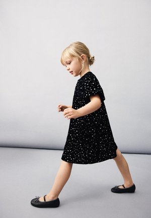 Mlada deklica z blond lasmi spetimi v figo, ki hodi, oblečena v črno obleko z belimi pikami in črne balerinke na enostavni ozadju.