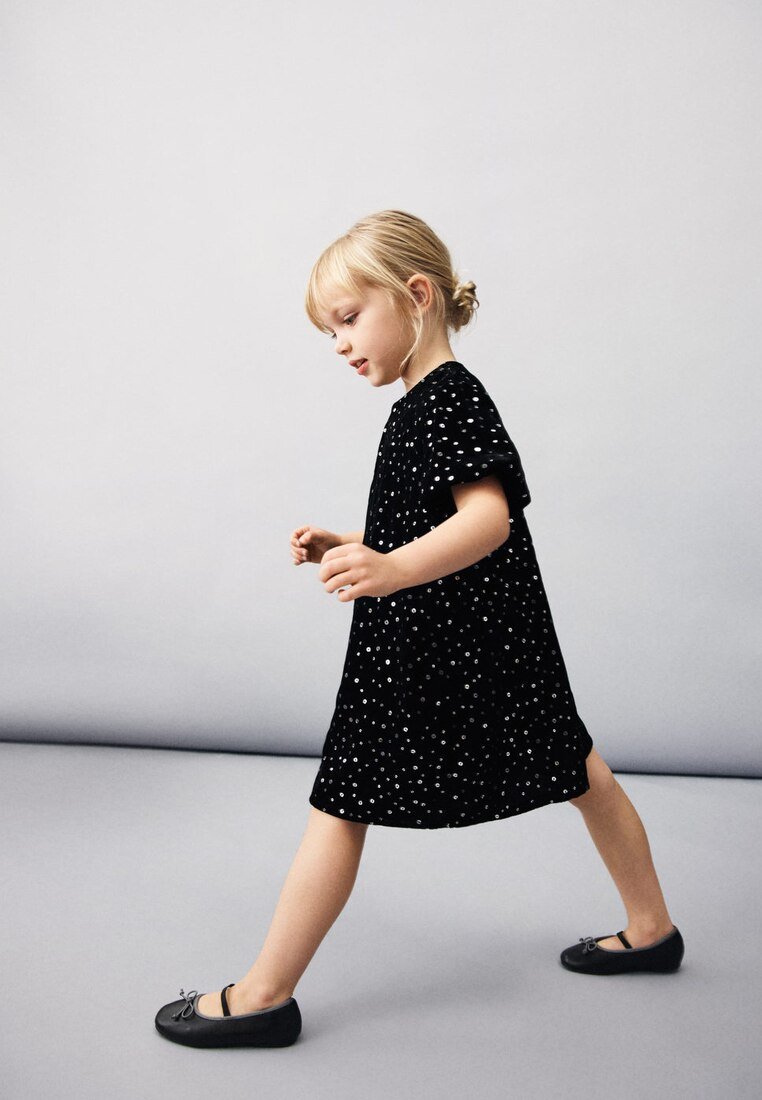 Mladá dívka s blond vlasy svázanými do drdolu, kráčející, oblečená v černých šatech s bílými puntíky a v černých balerínkách, na neutrálním pozadí.