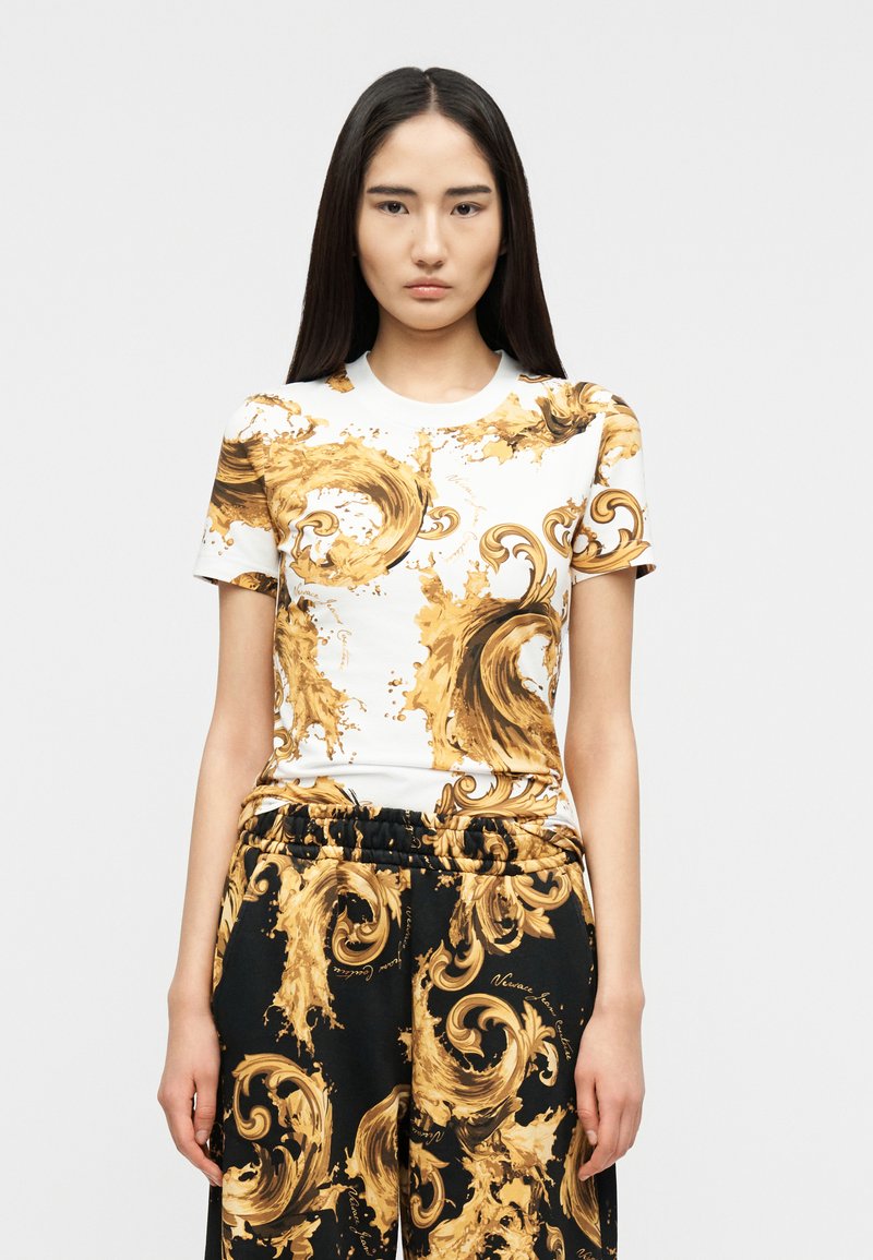 Γυναίκα με μακριά σκούρα μαλλιά, φορώντας ένα ταιριαστό λευκό και μαύρο σύνολο με μεγάλα χρυσά διακοσμητικά μοτίβα στροβίλου, στέκεται μπροστά σε απλό φόντο.