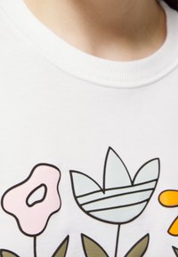 Vit T-shirt med blommig grafik. Har rosa, blåa och orange blommor med svarta konturer, gröna blad och ribbat nackband.
