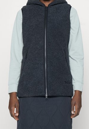 Osoba ubrana w czarną polarową kamizelkę na zamek błyskawiczny, nałożoną na jasnoniebieską koszulę z długim rękawem oraz ciemną pikowaną spódnicę, ukazana od szyi do połowy ud.