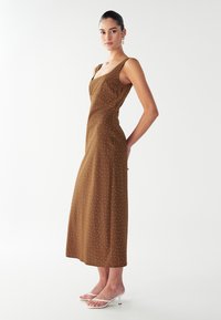 Joven mujer de pie vestida con un vestido midi marrón sin mangas con pequeños lunares blancos y sandalias de tacón blancas, frente a un fondo liso.