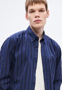 Tumesinise värvusega triibuline nööbitav särk tumedate siniste vertikaalsete triipudega, krae ja kaubamärgiga detailiga; kantud valge ümara kaelusega t-särgi peal.