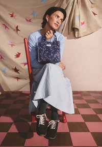 Nő piros széken ülve, világoskék ruhát, fekete sportcipőt és sötét zoknit viselve, egy kis tengerészkék virágos kézitáskát tartva, csillagmintás háttér előtt.