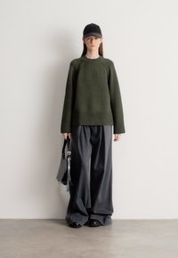 Oliviroheline soonikkoega kampsun koos soonilise äärega, kombineeritud laia halli pükstega. Modell hoiab musta kotti ja kannab musta mütsi. Lihtne taust.