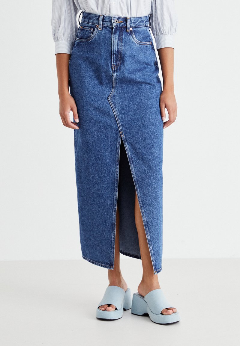 Jupe en denim bleu longue avec une fente à l'avant, taille haute et passants de ceinture. Associée à des sandales à plateforme bleu clair, mettant en valeur un style décontracté.