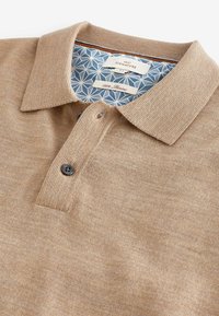 Beige polo paita, joka on valmistettu 100 % merinovillasta. Siinä on ribbireunus, kaksi mustaa nappia ja kuvioitu sininen sisävuori.
