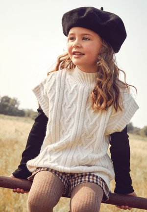 Cremfarbener, kabelgestrickter Pullover mit kurzen Ärmeln, über einem schwarzen Langarmshirt getragen. Braune karierten Shorts und strukturierte Strumpfhosen. Schwarze Baskenmütze setzt Akzente.