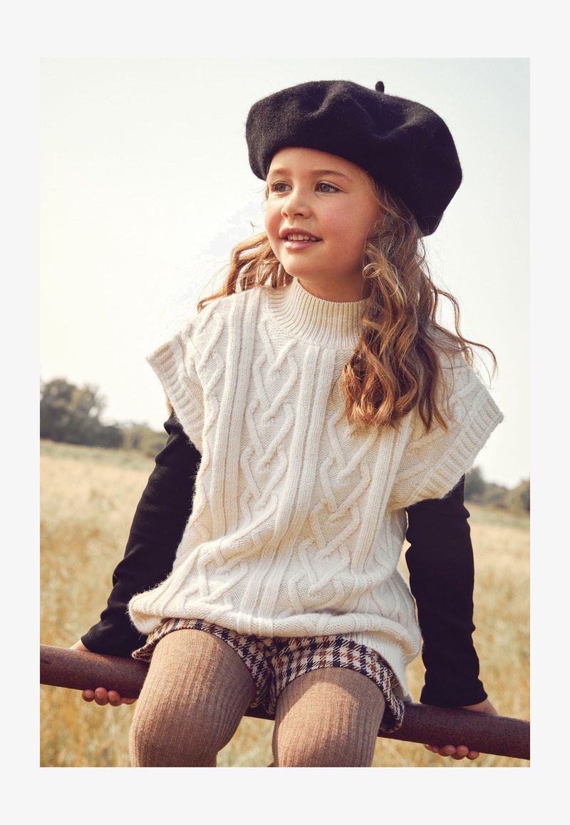 Maglione crema a trecce con maniche corte, indossato sopra una maglietta nera a maniche lunghe. Pantaloni corti a scacchi marroni e collant strutturati. Accenti in berretto nero.