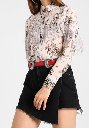 Blouse florale rose clair avec détails en franges, associée à une mini-jupe en denim noir. La tenue est complétée par une large ceinture rouge avec une boucle argentée.