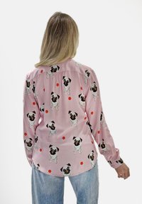 Rosa Bluse mit einem wiederholten Muster aus Mops-Hunden und roten geometrischen Formen. Verfügt über lange Ärmel, einen Kragen und einen Knopfverschluss. Glatte Textur.