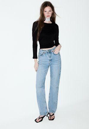 Junge Frau trägt schwarzen langärmeligen Croptop, hellblaue hoch taillierte Jeans, gepunktetes Halstuch und schwarze offene Absatzschuhe und steht.