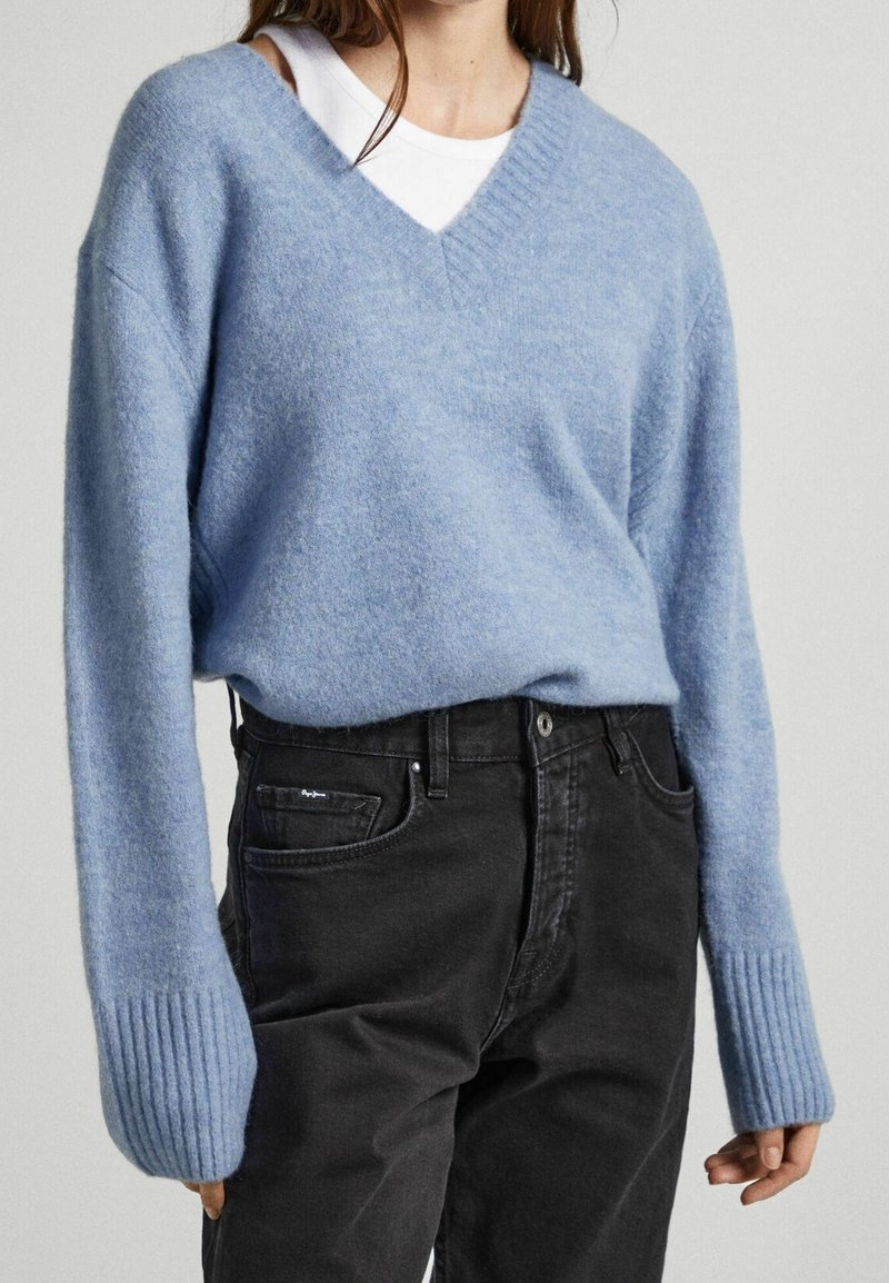 Femme portant un pull bleu clair à col en V et manches longues par-dessus un top blanc, associé à un jean noir taille haute sur un fond uni.