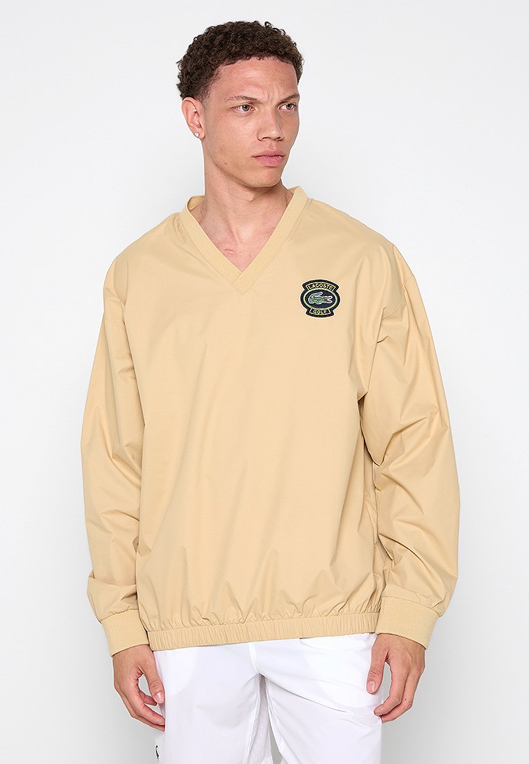 Veste pull beige avec un col en V, des poignets élastiques et un ourlet. Présente un logo crocodile noir brodé sur la poitrine. Tissu lisse et léger.