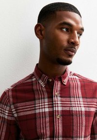 Joven con una camisa de cuadros rojos mira pensativamente hacia un lado contra una pared neutral, mostrando un estilo casual.