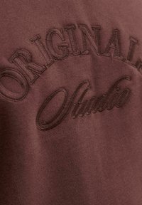 Brown fabric with raised embroidered text "ORIGINAL" and "Olivato" in different font styles on a garment.