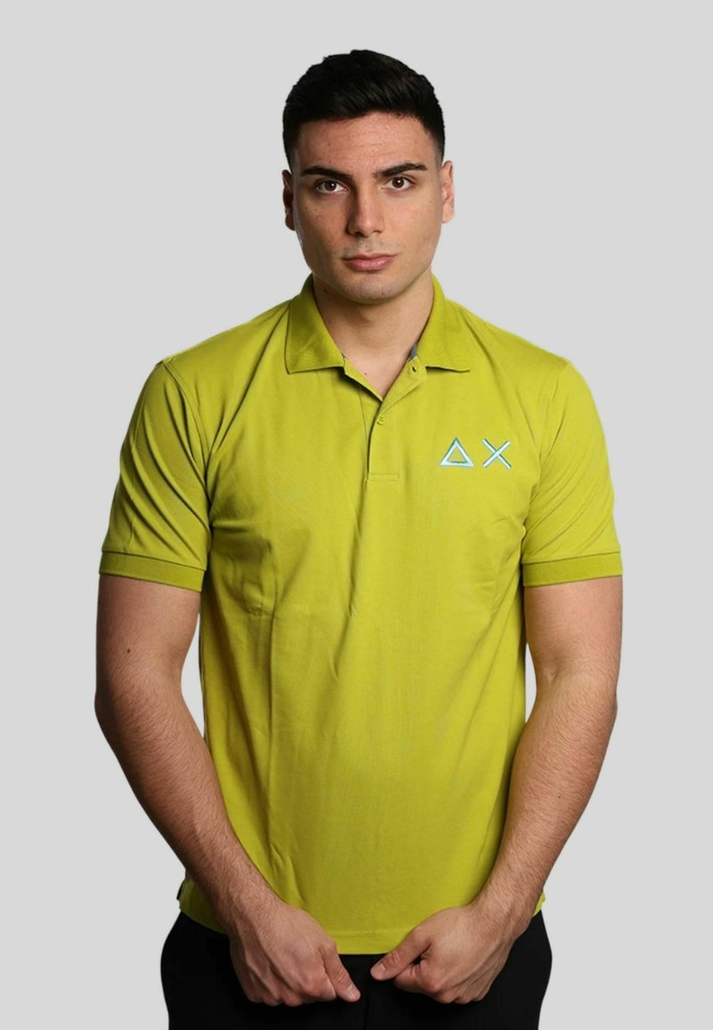 Polo di colore verde lime con colletto, maniche corte e un logo geometrico blu sul lato sinistro del petto. Tessuto liscio, design aderente.