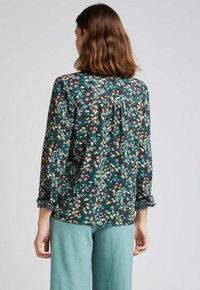 Blouse à manches longues florale en bleu sarcelle foncé avec des fleurs roses et jaunes. Dispose d'un détail froncé au dos et de poignets roulés. Portée avec un pantalon bleu sarcelle clair.