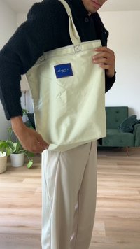 Krämfärgad tote-väska med blå GANT-logotyp. Tillverkad av ett slätt material, med långa handtag och synlig sömnad. Enkel, rektangulär form.