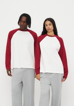Due persone sono in piedi fianco a fianco indossando magliette a maniche lunghe bianche e rosse abbinate con pantaloni grigi su uno sfondo semplice.