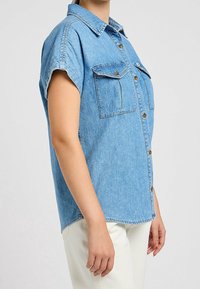 Blue denim short-sleeve shirt with a button-down front, two chest pockets, and a rounded hemline; light blue wash with faded texture.