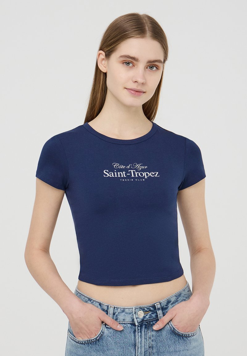 T-shirt cropped blu navy con maniche corte, con testo stampato bianco "Côte d'Azur Saint-Tropez Tennis Club" sul petto.