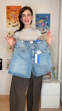 Γυναίκα με μακριά σκούρα μαλλιά που κρατάει ανοιχτόχρωμα τζιν σορτς με ετικέτες, στέκεται μπροστά σε έναν τοίχο με πολύχρωμες αφίσες κινουμένων σχεδίων.