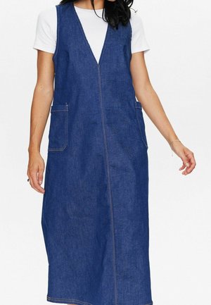 Femme portant une robe en jean bleue sans manches avec deux poches avant par-dessus une chemise blanche à manches courtes, debout devant un fond blanc.