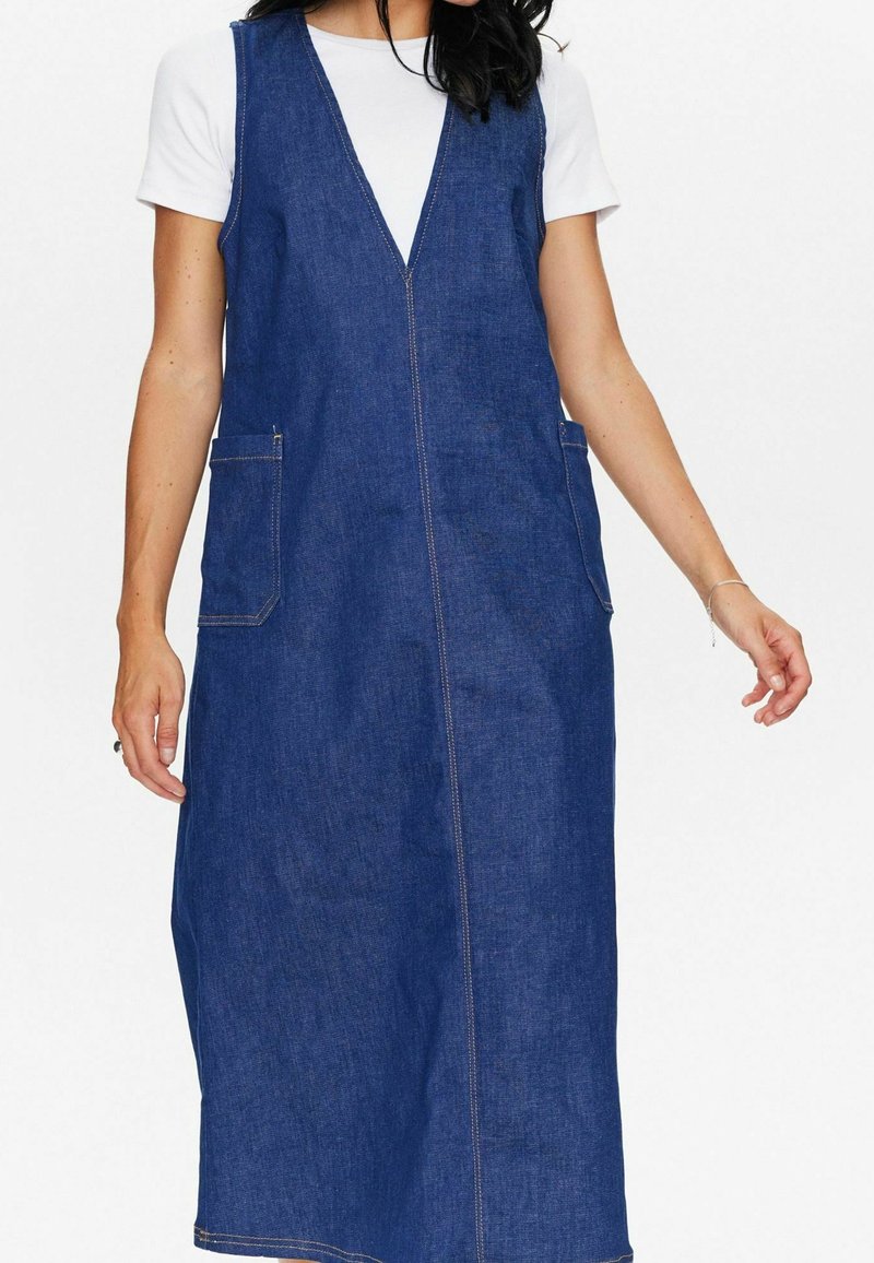Femme portant une robe en jean bleue sans manches avec deux poches avant par-dessus une chemise blanche à manches courtes, debout devant un fond blanc.