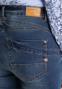Jean en denim de couleur bleu foncé avec une étiquette en cuir portant le mot "cream". Les caractéristiques comprennent des coutures contrastantes et un détail de bouton en métal décoratif.