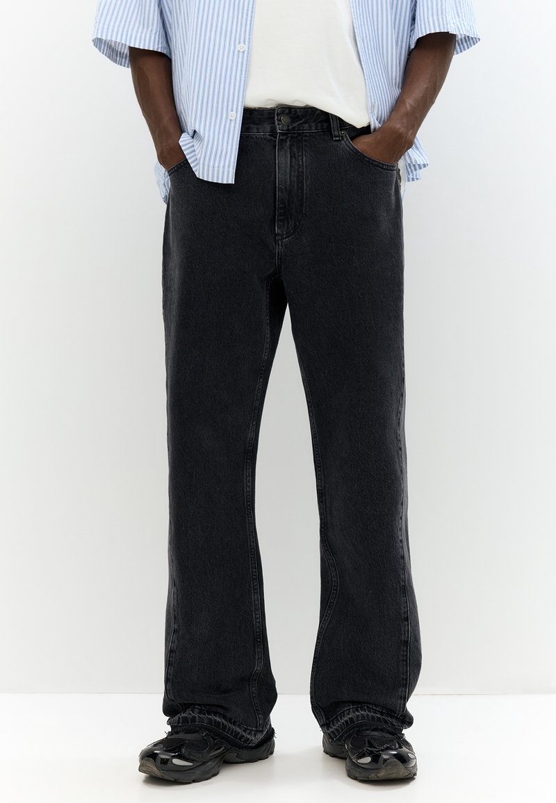 Personne portant un jean noir ample, des baskets noires, une chemise blanche et une chemise à manches courtes rayée bleu et blanc, les mains dans les poches.