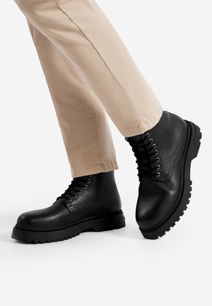Czarne skórzane buty z teksturowanym wykończeniem, sznurowane, z masywną czarną podeszwą i wzmocnionym noskiem. Sparowane z beżowymi spodniami.
