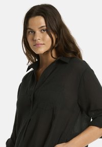 Una giovane donna con capelli lunghi e ondulati posa indossando una camicia nera con bottoni, guardando direttamente in camera con un'espressione neutra.