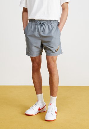 Shorts de sport gris avec une taille élastique et un cordon de serrage, présentant un logo latéral en orange. Portés avec des baskets blanches et des chaussettes de longueur crew.