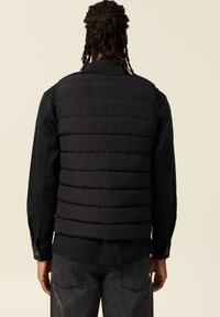 Chaleco acolchado negro con un patrón de acolchado vertical, llevado sobre una camisa negra de manga larga. La textura es suave con un acabado mate.