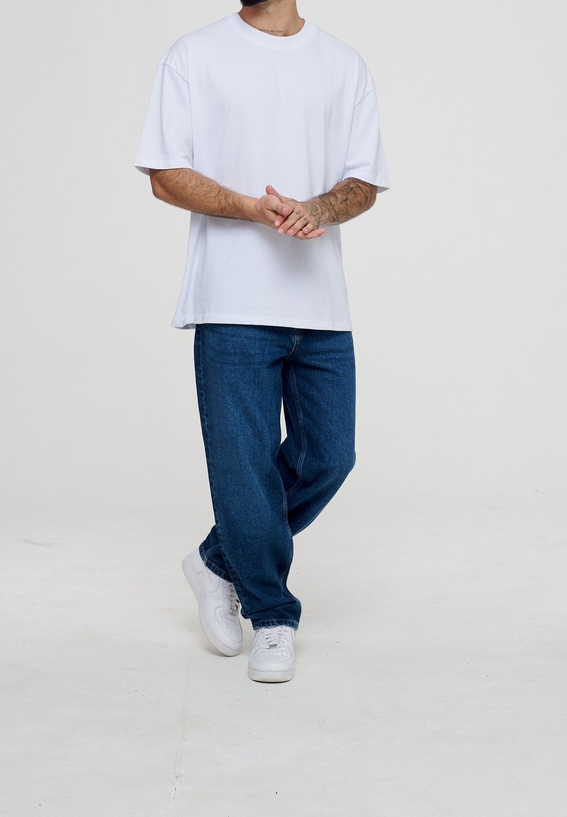 Homme portant un T-shirt blanc oversize, un jean bleu et des baskets blanches, avec les bras tatoués et les mains jointes devant, debout sur un sol blanc.
