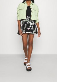 Veste courte vert clair avec deux poches avant, portée sur un haut noir et une jupe noire ornée d'un motif de feuilles et de fleurs tropicales. Sandales blanches.