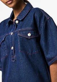 Tumšas krāsas džinsu krekls ar pogu slēdzēju, īsām piedurknēm, krūšu kabatām un kontrastējošām sarkanām šuvēm. Gluda tekstūra, ikdienišķs dizains.