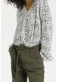 Weiße Bluse mit schwarzem gesprenkeltem Muster, V-Ausschnitt; kombiniert mit olivgrünen Hosen, betont durch einen gebundenen Bund und seitliche Taschen.