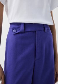 Pantaloni su misura viola con una texture liscia, caratterizzati da una chiusura a bottone visibile e un cinturino ben cucito, abbinati a una maglietta bianca.