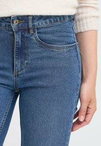 Moteris vilkinti mėlynus džinsus ir kreminę megztinę, ranka atsirėmusi šalia, priartinta prie juosmens ir viršutinio šlaunų ploto.