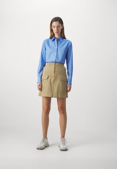 Camicia azzurra chiara con colletto, abbinata a una gonna cargo beige con tasche frontali, indossata con sneakers grigie e bianche.